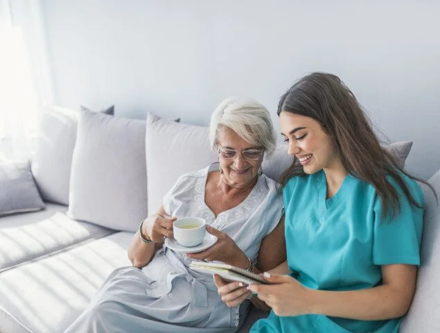 Happy patient is holding caregiver for a hand while spending time together. Elderly woman in nursing home and nurse. Aged elegant woman and tea time at nursing home