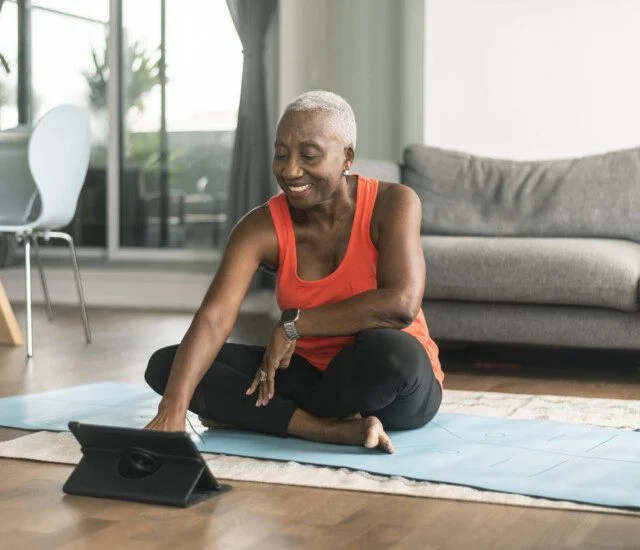 A beautiful senior woman takes an online yoga class. She is interacting with the teacher on her tablet and about to commence her class. She is wearing casual active wear and is taking the class in her lounge room. She is sitting on a blue yoga mat.