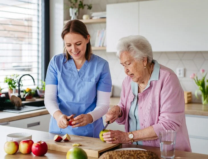 Home caregiver and elderly woman baking apple pie together, cutting apples. Household chores for senior woman, help from nurse.