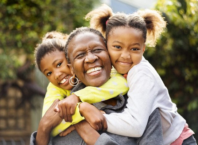senior woman seated outdoors with her two grandchildren, who are embracing her from behind and smiling