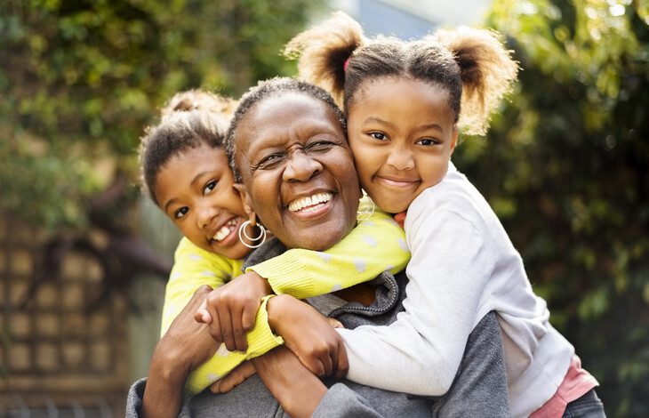 senior woman seated outdoors with her two grandchildren, who are embracing her from behind and smiling