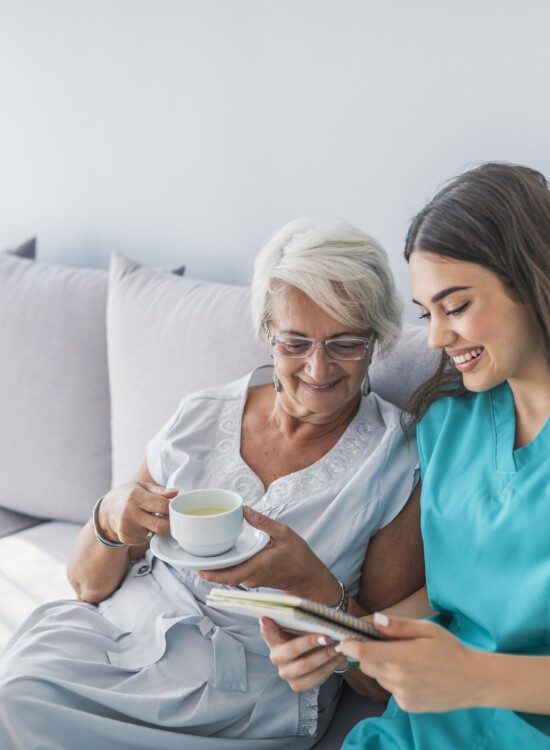 Happy patient is holding caregiver for a hand while spending time together. Elderly woman in nursing home and nurse. Aged elegant woman and tea time at nursing home